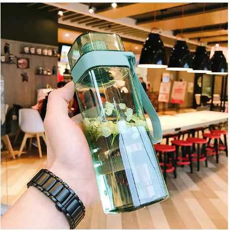 Hand holding a trendy square water bottle with a blue strap in a modern cafe setting