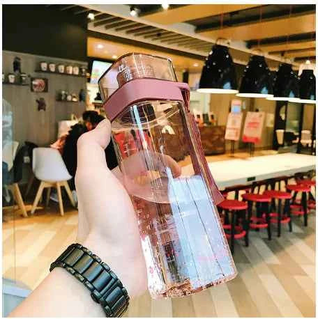 Hand holding a trendy square water bottle with pink lid in a modern cafe setting