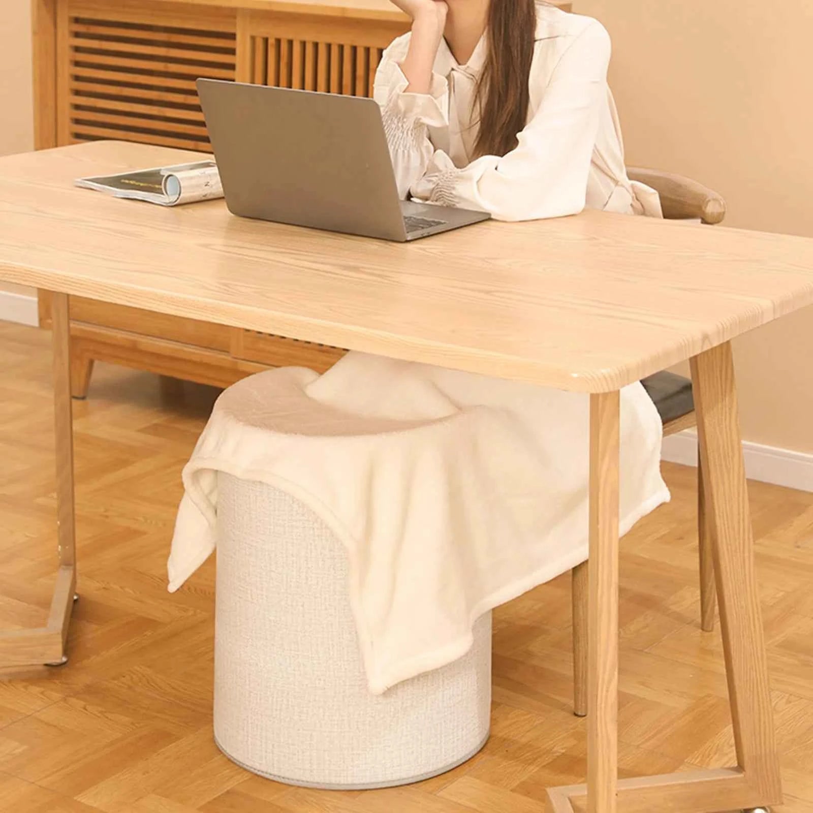 Woman using portable heated foot warmer for home under wooden desk while working on laptop