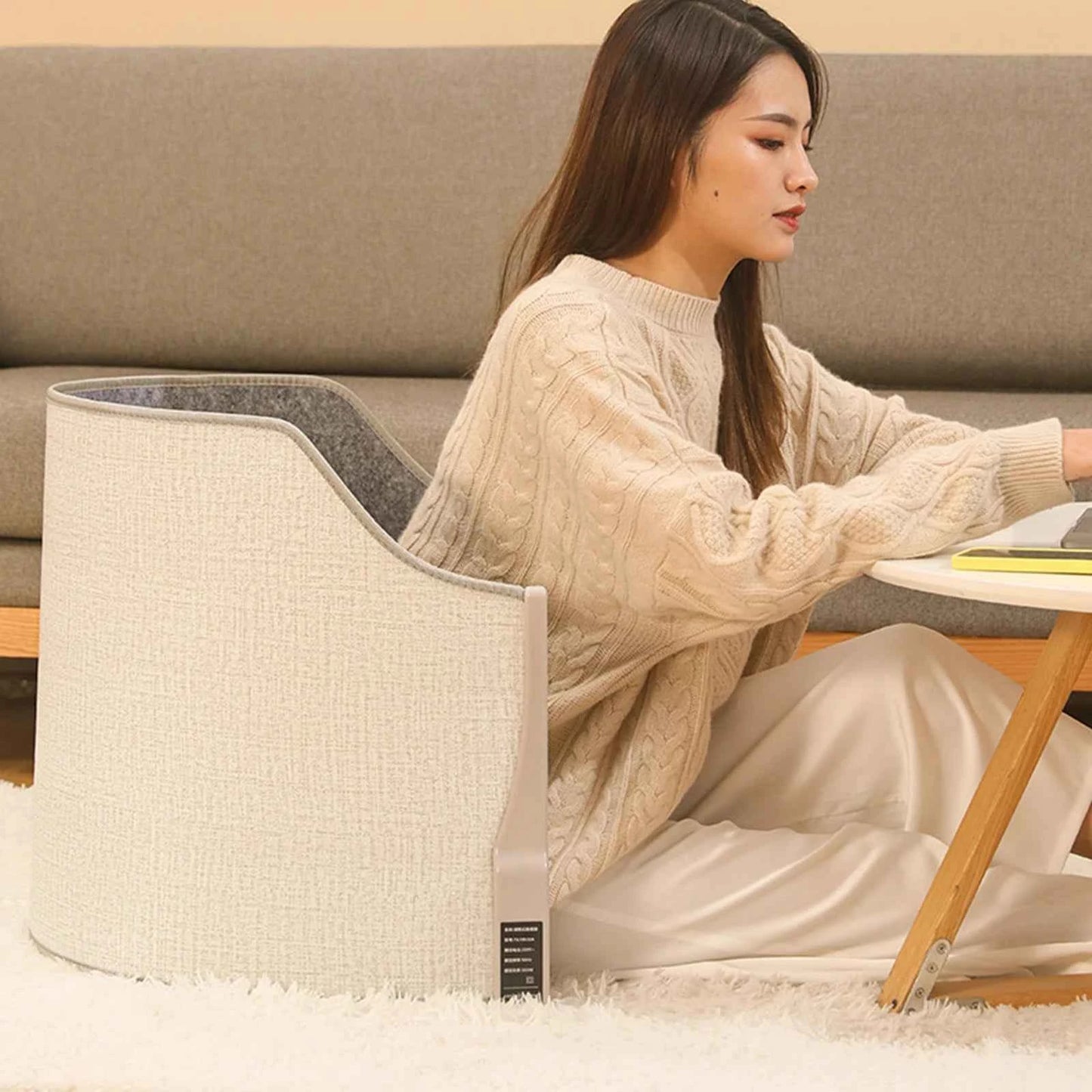 Woman using a portable heated foot warmer for home while sitting on the floor by a wooden table